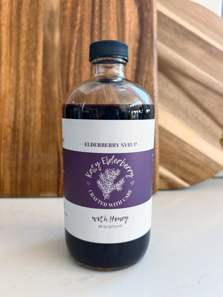 Katy Elderberry organic elderberry syrup with raw honey, 16 oz amber glass bottle, small-batch herbal immune support remedy on a marble countertop with wooden cutting boards.