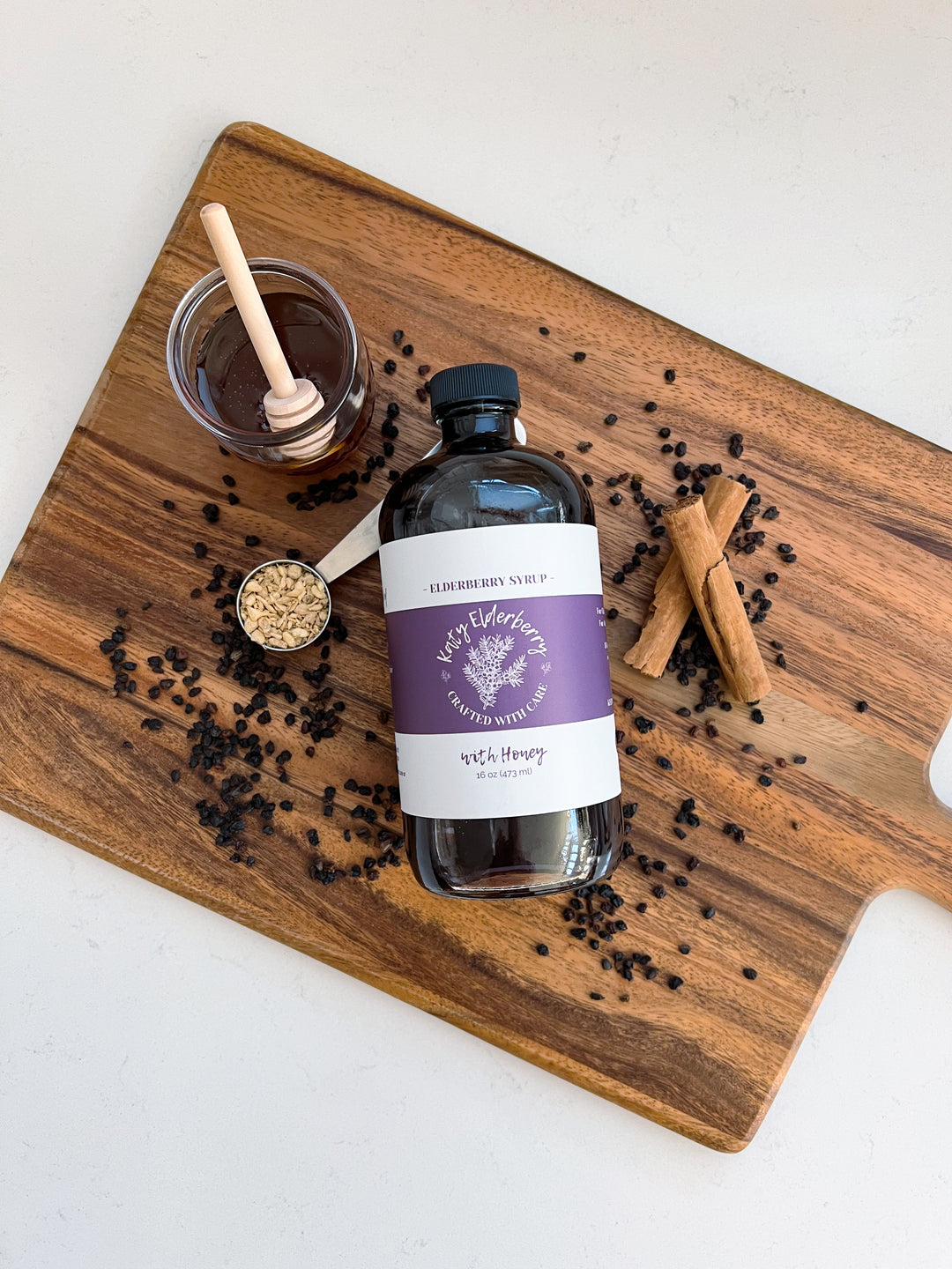 Katy Elderberry organic elderberry syrup with raw honey, a small-batch immune support remedy, displayed in an amber glass bottle on a wooden cutting board with cinnamon sticks, dried elderberries, and a jar of honey.