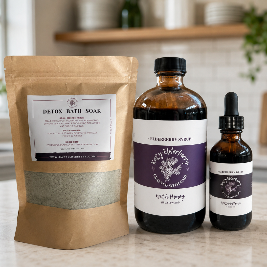 Three wellness products from Katy Elderberry arranged on a kitchen counter: a kraft paper stand-up pouch labeled Detox Bath Soak, an amber bottle labeled Elderberry Syrup, and a smaller amber dropper bottle labeled Wellness to go.